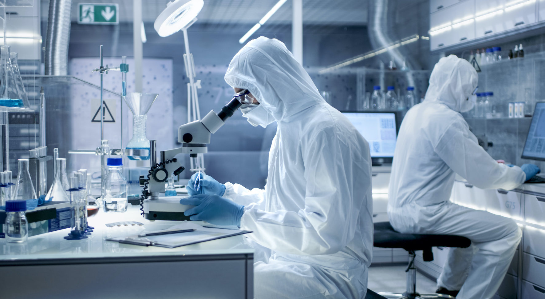 In a Secure High Level Laboratory Scientists in a Coverall Conducting a Research. Biologist Adjusts Samples in a Petri Dish with Pincers and Examines Them Under Microscope and His Colleague Analyzes Results on a Computer.