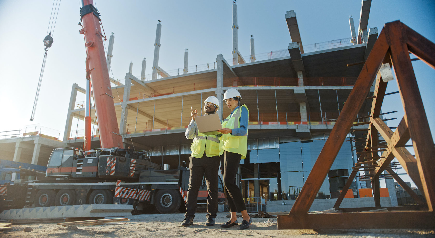 Two Specialists Inspect Commercial, Industrial Building Construction Site. Real Estate Project with Civil Engineer, Investor Use Laptop. In the Background Crane, Skyscraper Concrete Formwork Frames