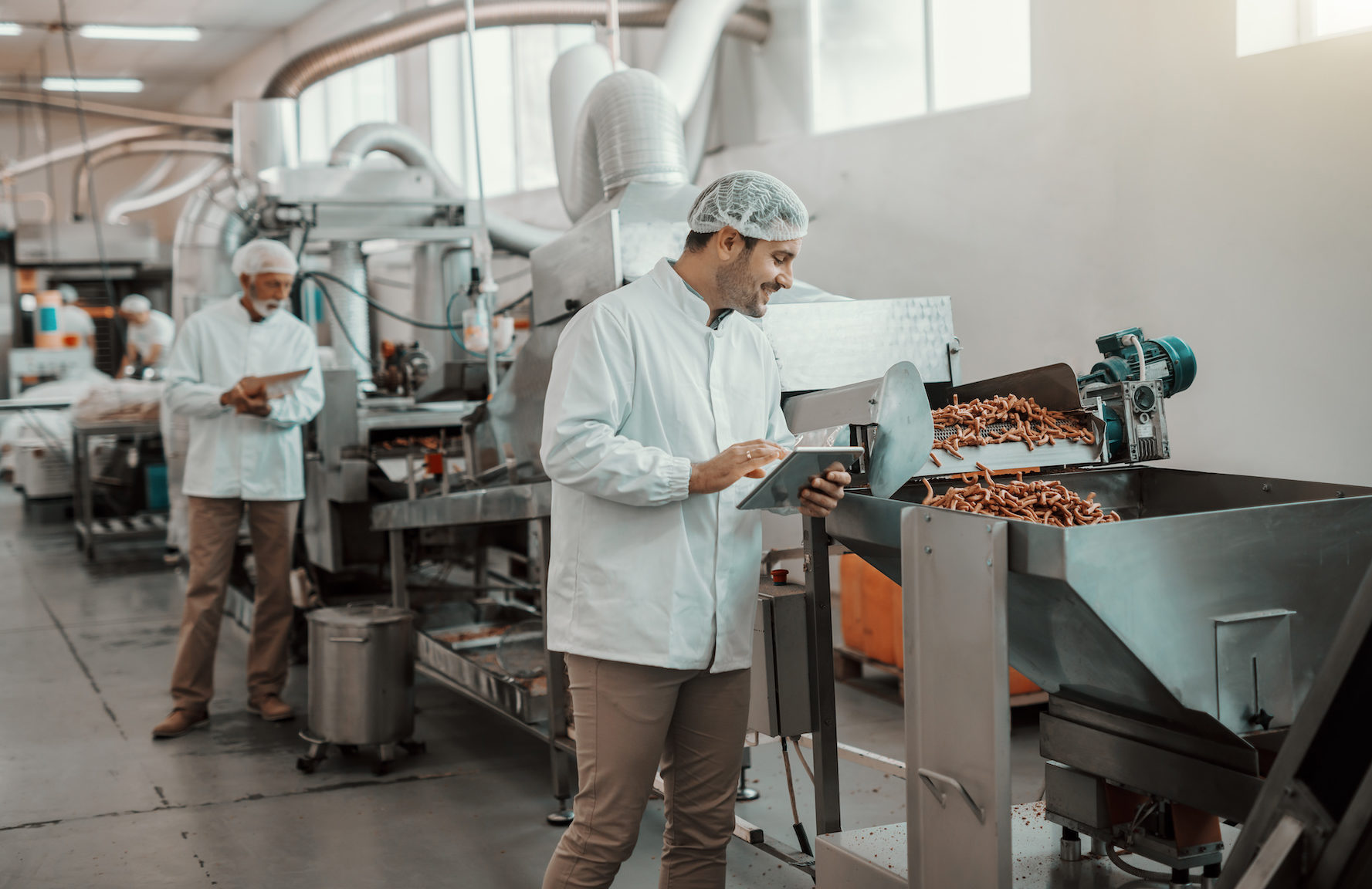 Supervisor evaluating quality of food in food plant while holding tablet. Man is dressed in white uniform and having hair net.
