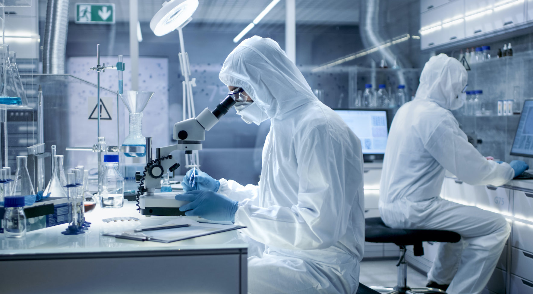 In a Secure High Level Laboratory Scientists in a Coverall Conducting a Research. Biologist Adjusts Samples in a Petri Dish with Pincers and Examines Them Under Microscope and His Colleague Analyzes Results on a Computer.