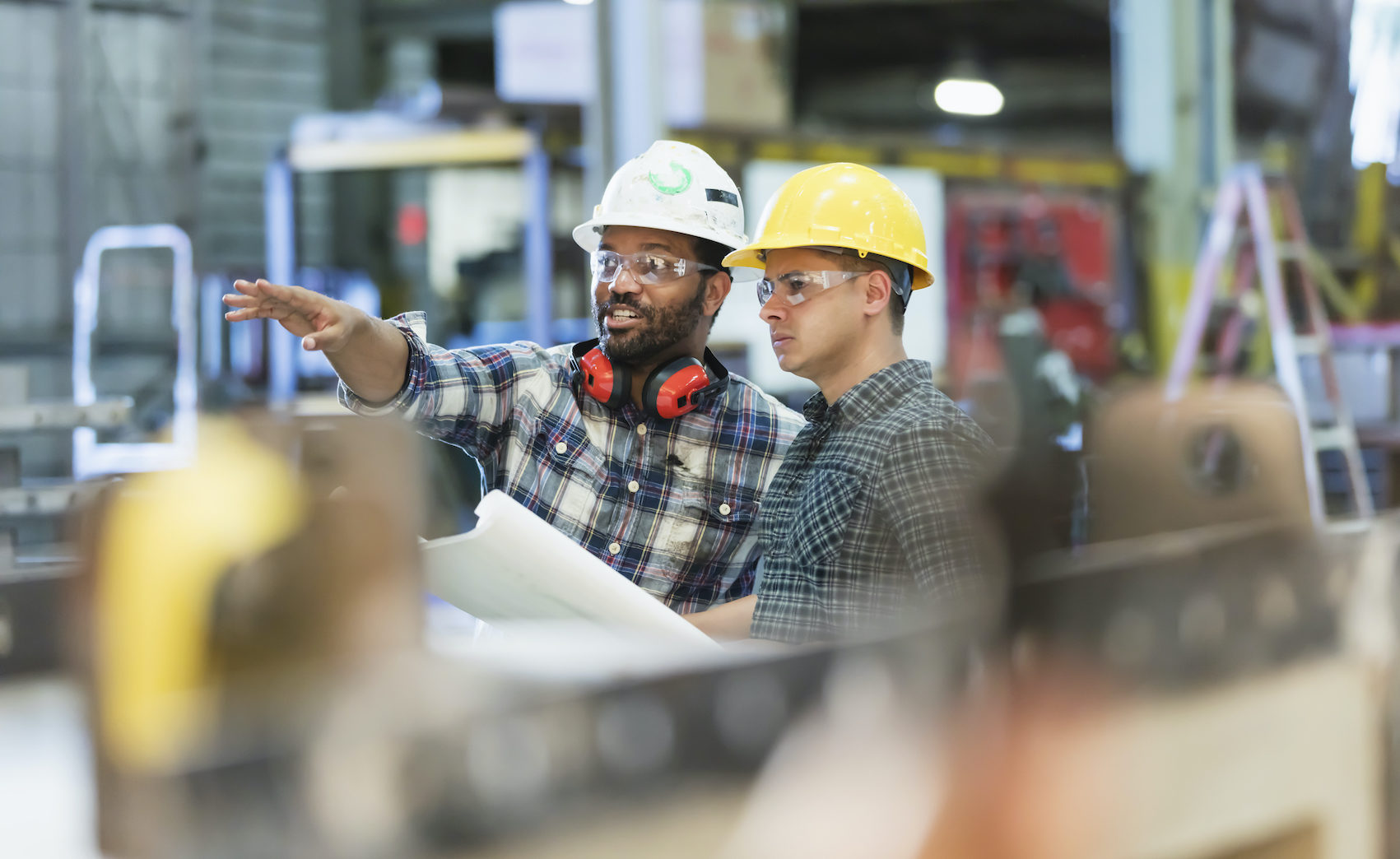 Two workers talking in a metal fabrication plant wearing hardhats and protective eyewear.