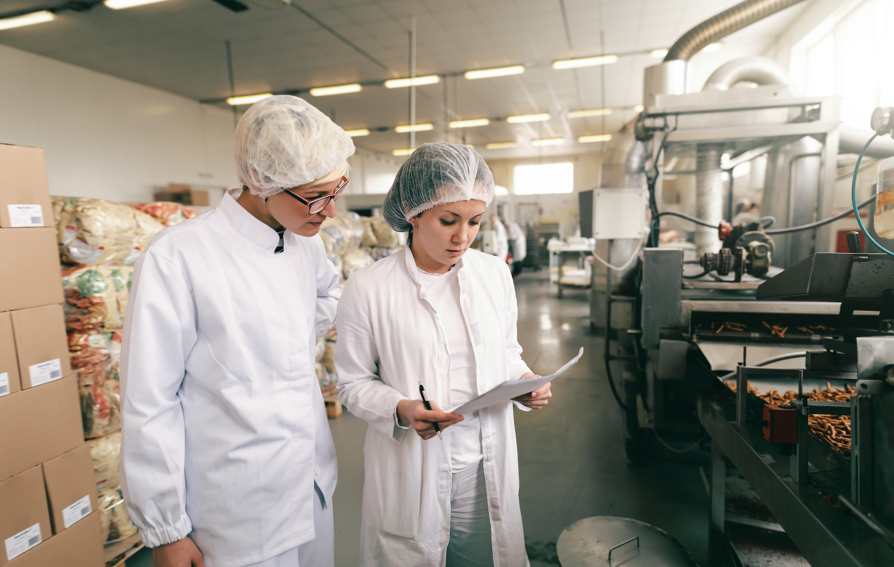 Two quality professionals in white sterile uniforms checking quality of salt sticks while standing in food factory.