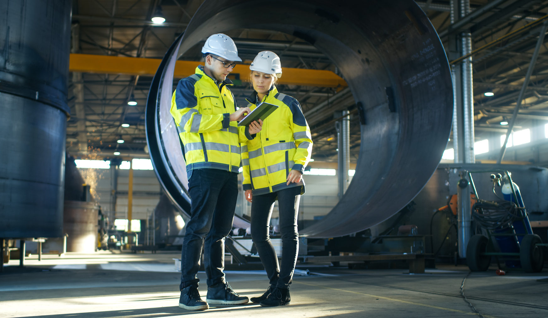 Industrial Engineers in Hard Hats Discuss New Project while Using Tablet Computer. They're Making Calculated Engineering Decisions.They Work at the Heavy Industry Manufacturing Factory.