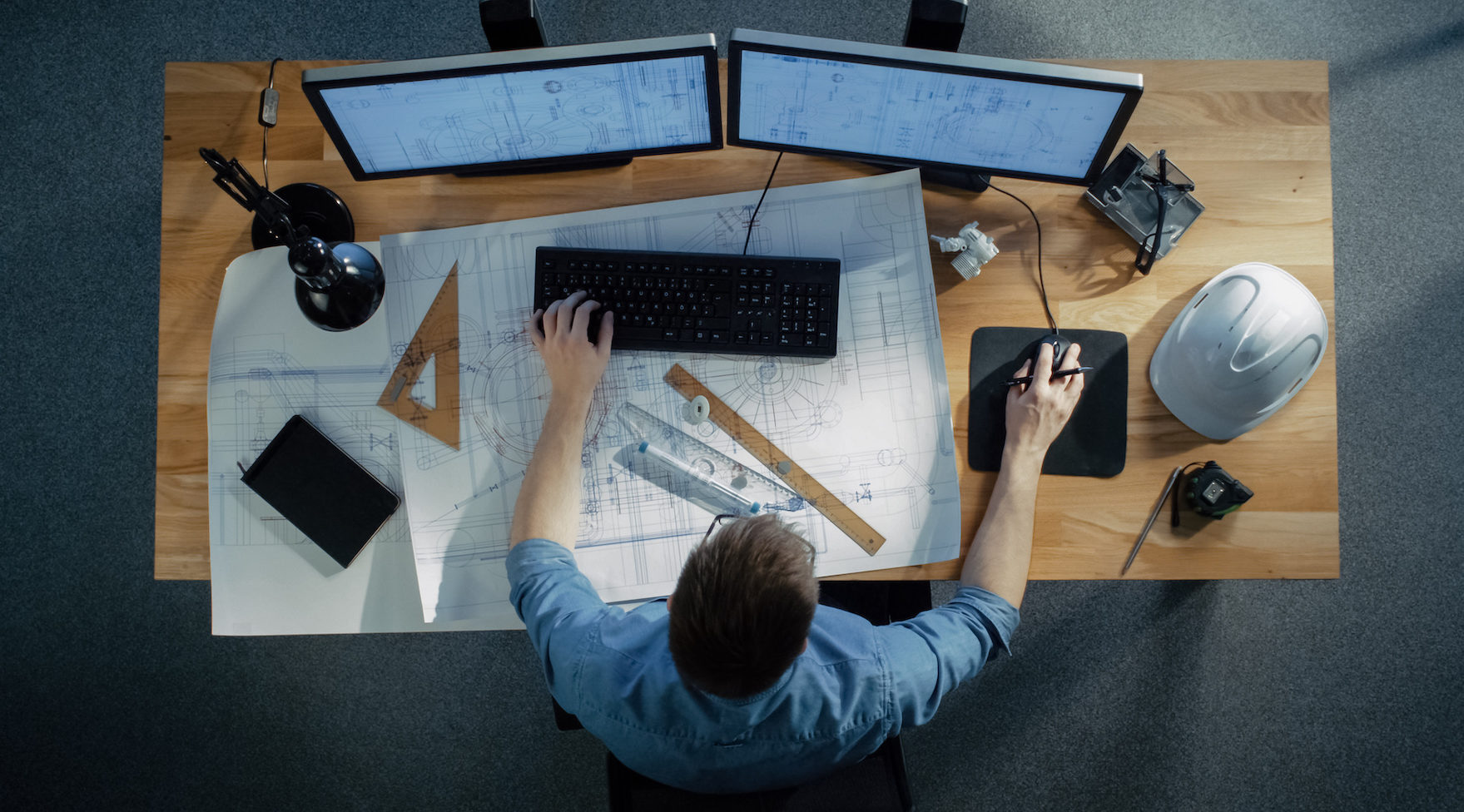 Top View of a Technical Engineer Working on His Blueprints, Drawing Plans, Using Desktop Computer. Various Useful Items Lying on his Table.